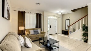 Una sala de estar con un sofá color beige, una mesa de centro de vidrio y una lámpara. Se observa una escalera con barandilla y una planta en maceta.