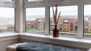A room with a bed, a window with a view of brick buildings, and a vase with reeds.