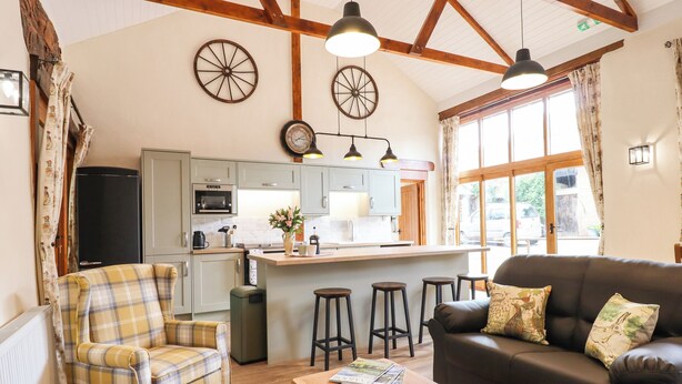 Una sala de estar acogedora con un sofá, un sillón a cuadros y una mesa de centro de madera. Cuenta con una zona de cocina que incluye un refrigerador, un microondas y una pequeña isla con taburetes. La habitación tiene vigas de madera en el techo y ventanas amplias.