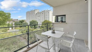 Ein Balkon mit weißem Tisch und Stühlen, einer Glaseinfriedung und einem dekorativen Gegenstand, sowie Blick auf umliegende Gebäude und Grünflächen.