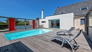 En modern utomhuspool med trädäck och solstolar, en röd accentvägg och en fyr i bakgrunden.