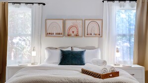 A neatly arranged bedroom with a large bed, a bedside table with a lamp, a nightstand, and a window with curtains. There are three framed artworks above the bed.