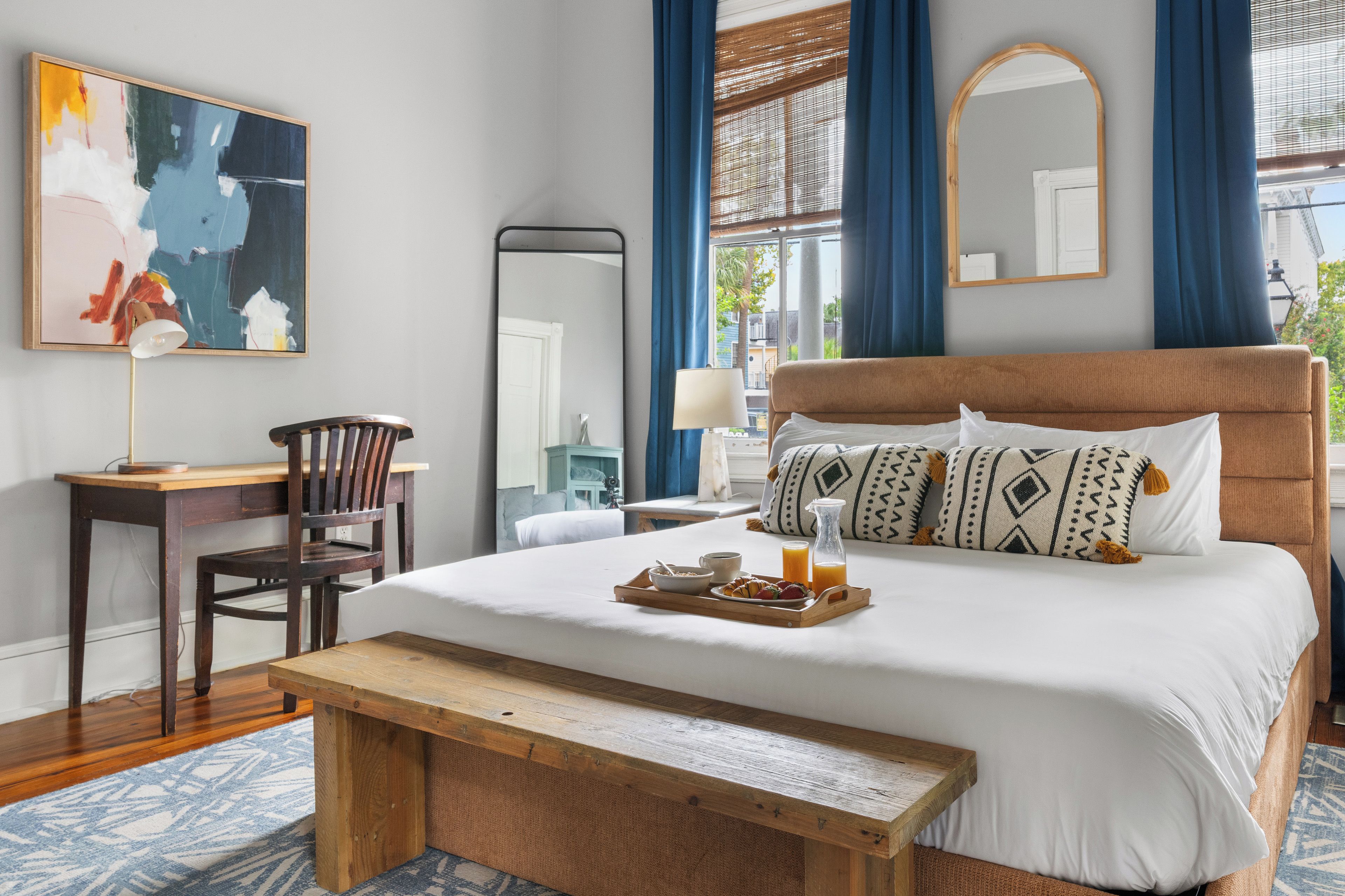 A hotel room with a bed, a desk, a chair, a bedside table, a mirror, and a window with curtains.