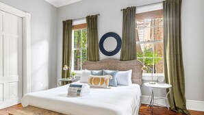 A bedroom with a large bed, a round mirror, a small side table, and a window with curtains.
