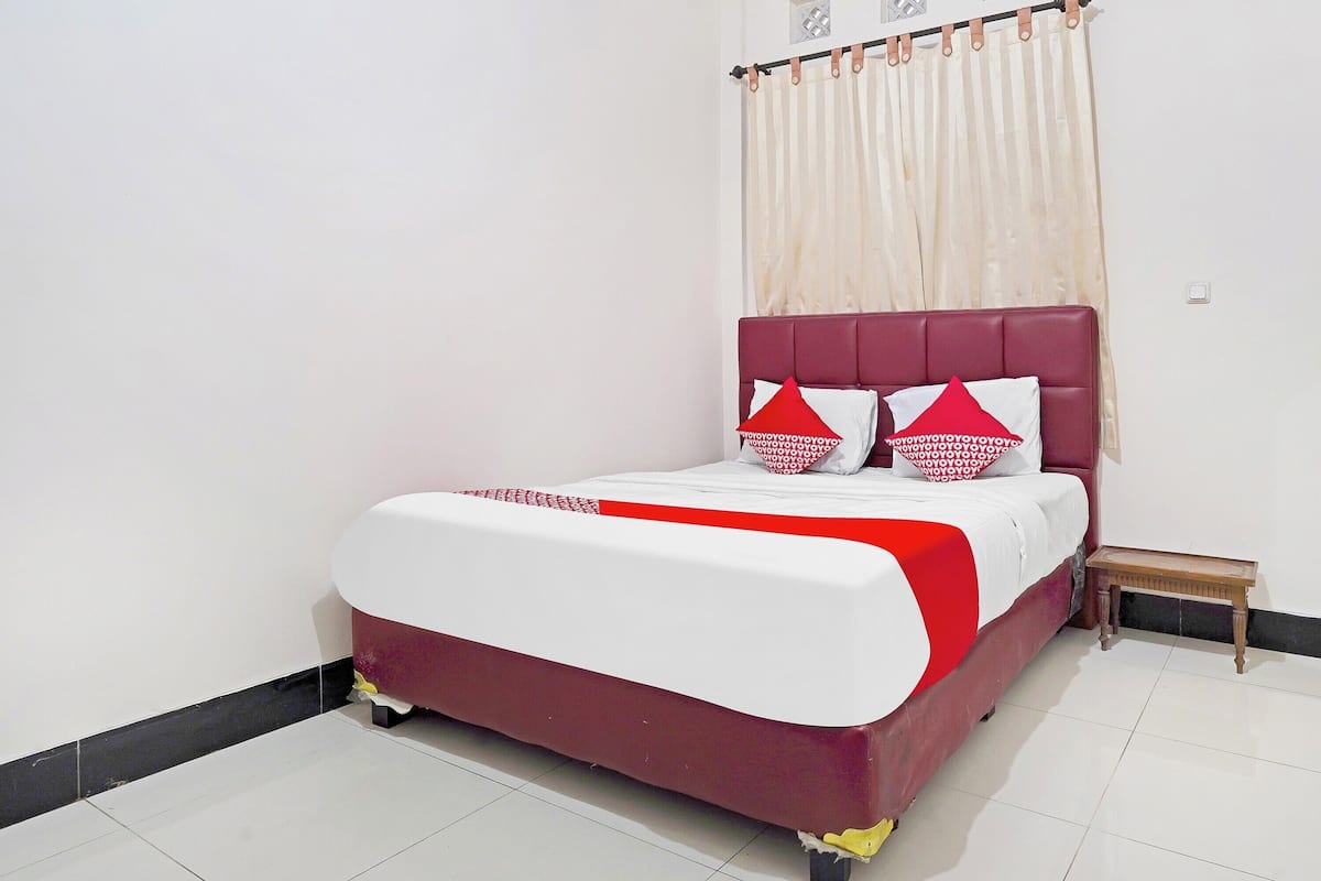 A neatly made bed with a red and white bedspread, a small wooden nightstand beside it, and a sheer white curtain covering the window.