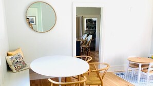Een kamer met een ronde witte tafel, vier rieten stoelen, een ronde spiegel en een witte bank met een gekleurd kussen.