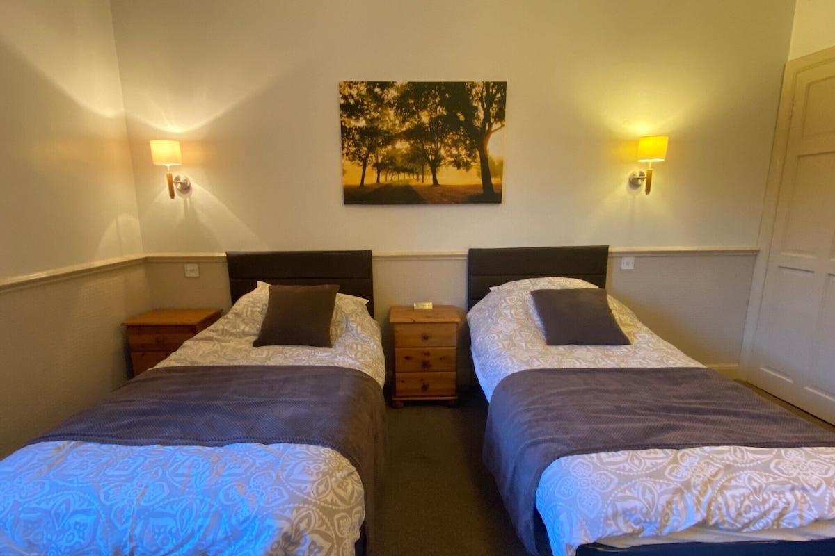 A double room with two beds, each with a headboard, bedside tables, and lamps. A painting of trees is on the wall above the beds.