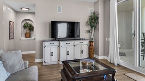 Een woonkamer met een flatscreen televisie, witte kasten, een glazen salontafel, een grijze bank en een schuifpui naar een balkon.