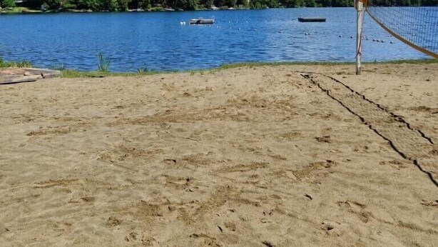 A beach volleyball court with a net, a sandy foreground, and a lake with boats and trees in the background. - 3 bedroom home steps away from beach/lake (Petersburg)