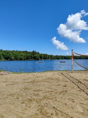 A beach volleyball court with a net, a sandy foreground, and a lake with boats and trees in the background.