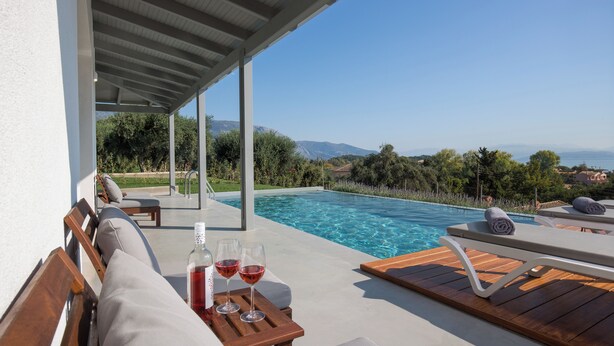 Uma área à beira da piscina com espreguiçadeiras, uma mesa com copos de vinho tinto e vista para o mar e montanhas.