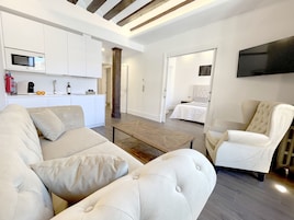 A modern living room with a sofa, armchair, and coffee table. There is a kitchen area with appliances and a dining table.