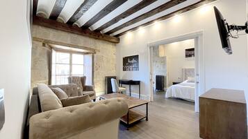 Un'ampia zona giorno con soffitto alto, travi a vista e una grande finestra. Si vedono un divano, un tavolino da caffè e una poltrona. Un'apertura conduce a una camera da letto con un letto e un comodino.