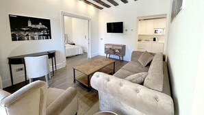 Un salon moderne avec un canapé, des fauteuils, une table basse et un téléviseur. On y trouve également un coin cuisine avec des placards et un four à micro-ondes. Un lit est visible au fond.
