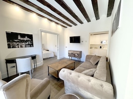 A modern living room with a sofa, armchairs, a coffee table, and a TV. There is a kitchen area with cabinets and a microwave. A bed is visible in the background.