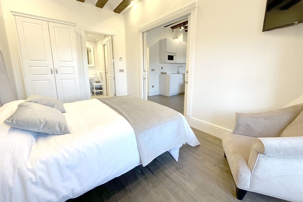 A bedroom with a bed, a chair, and a kitchenette in the background.