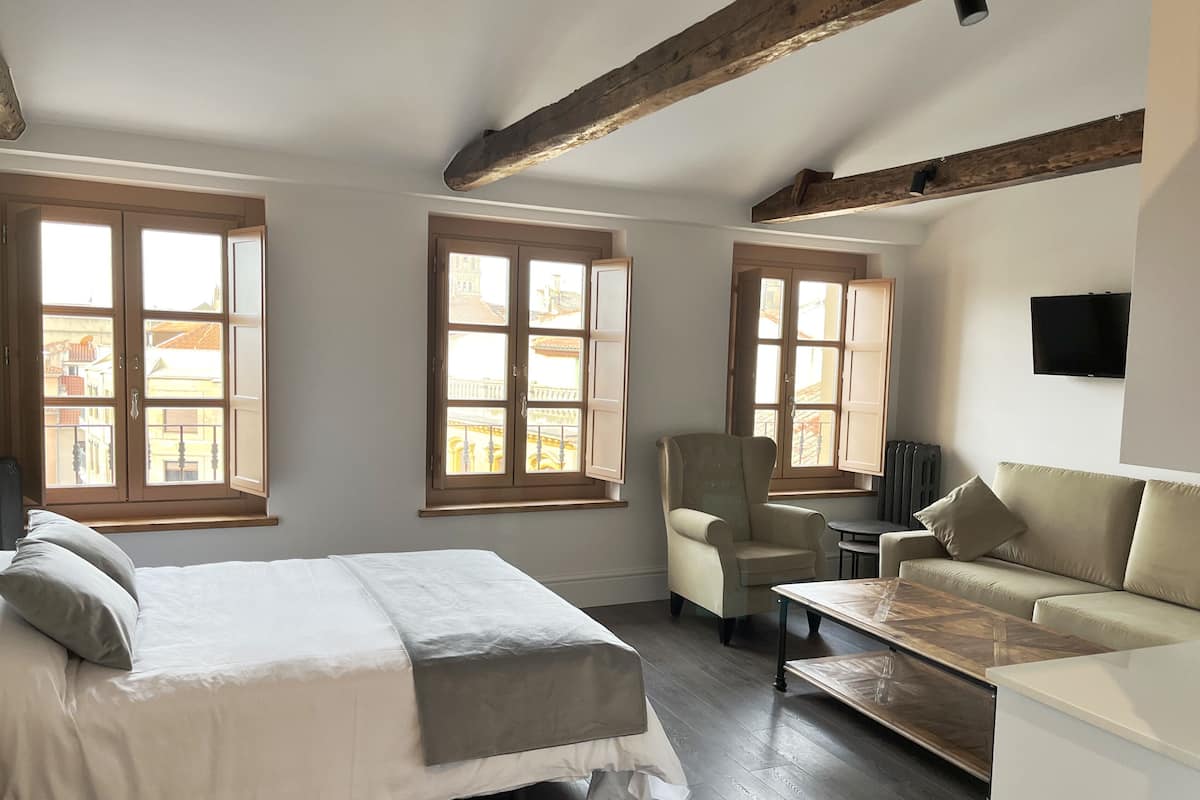 A room with a bed, a chair, a sofa, and a table. There are windows with wooden frames and a radiator on the wall.