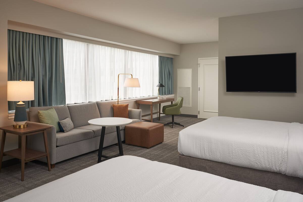 A hotel room with a sofa, two single beds, a desk with a chair, a lamp, and a TV.