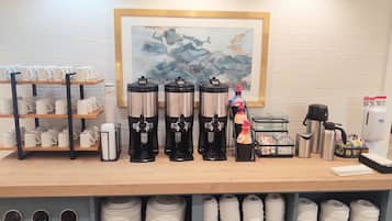 A coffee station with multiple coffee makers, a stack of disposable cups, and a framed painting on the wall.