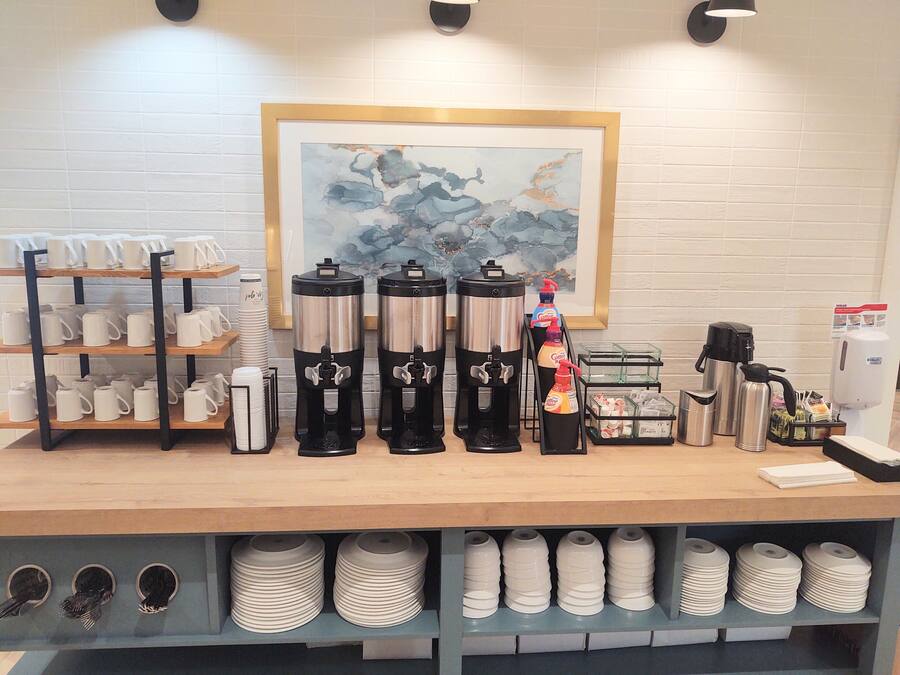 A coffee station with multiple coffee makers, a stack of disposable cups, and a framed painting on the wall.