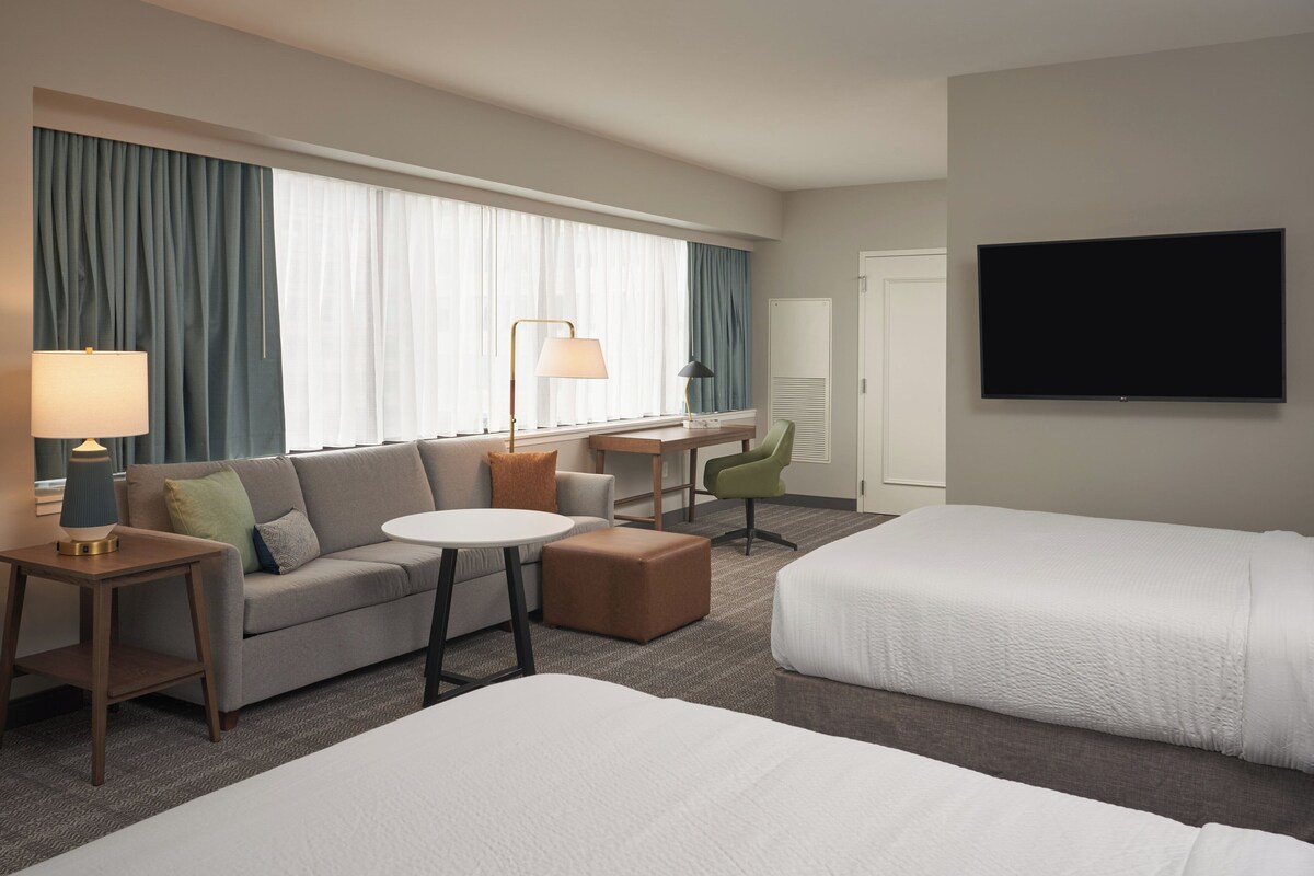 A hotel room with a sofa, two single beds, a desk with a chair, a lamp, and a TV.