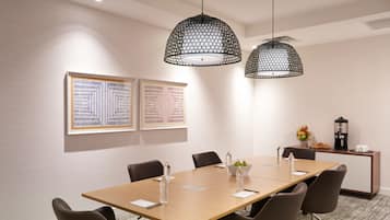 A conference room with a long table, six chairs, two pendant lights, framed artwork on the wall, and a coffee station.