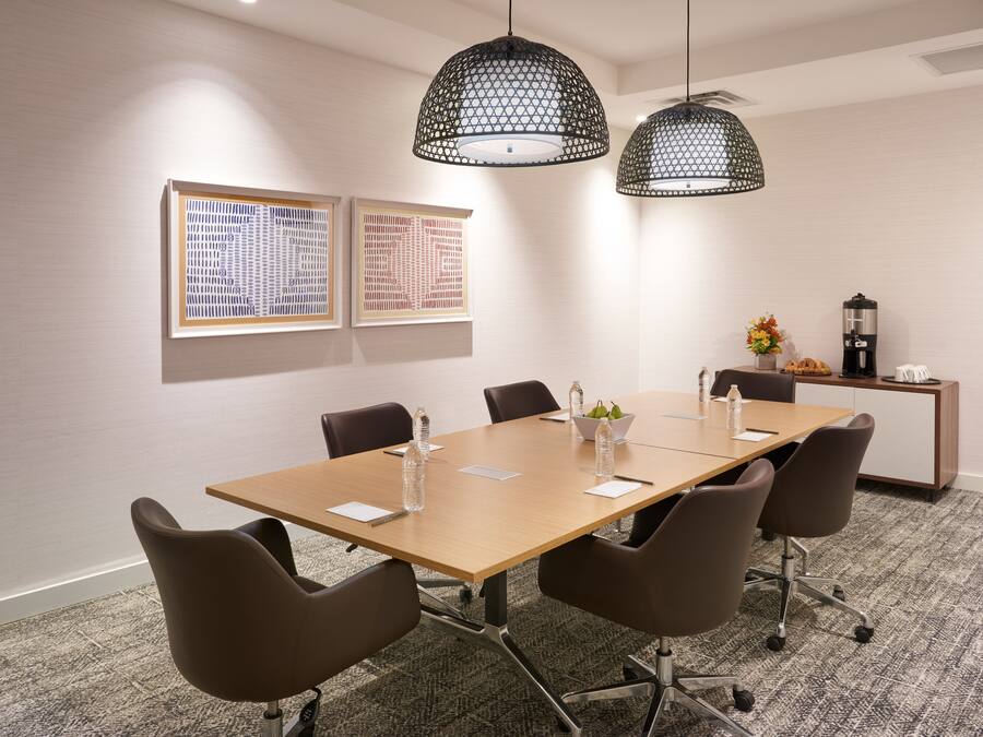 A conference room with a long table, six chairs, two pendant lights, framed artwork on the wall, and a coffee station.