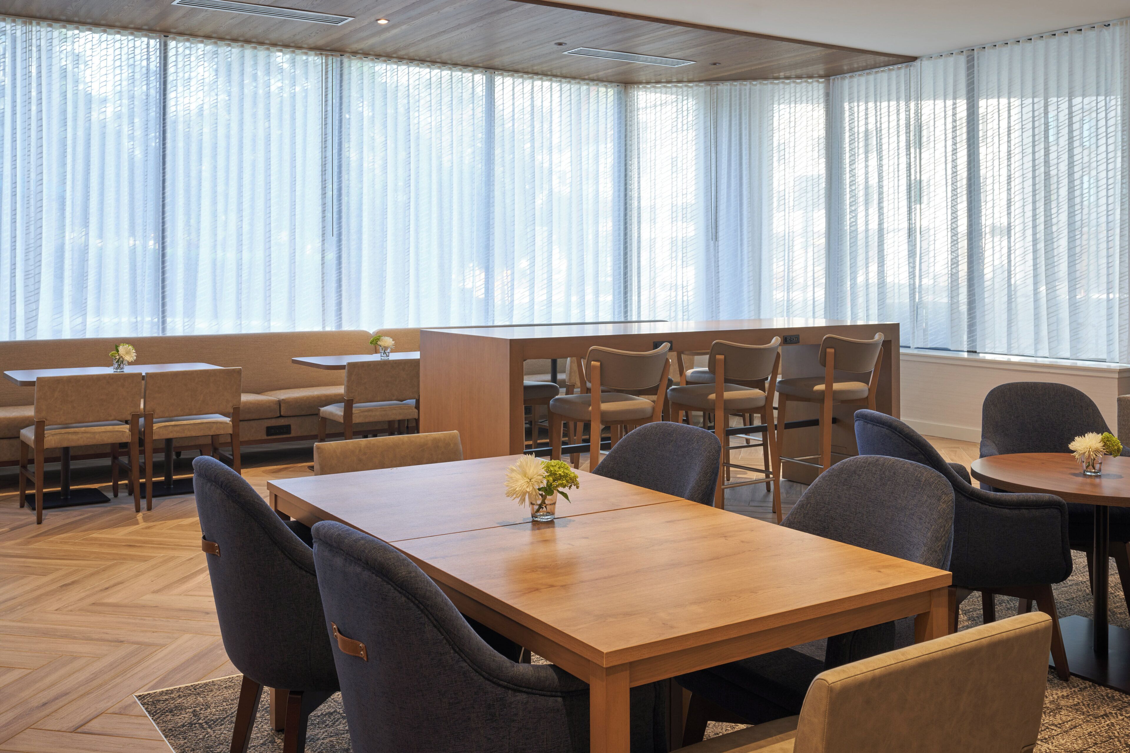 A dining area with multiple tables and chairs, large windows with sheer curtains, and a herringbone wood floor.