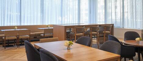 A dining area with multiple tables and chairs, large windows with sheer curtains, and a herringbone wood floor.
