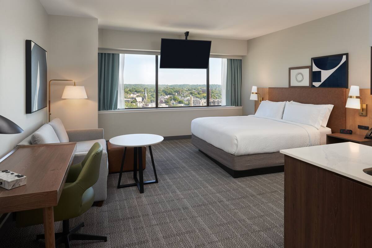A hotel room with a bed, a desk, a chair, a small table, and a TV.