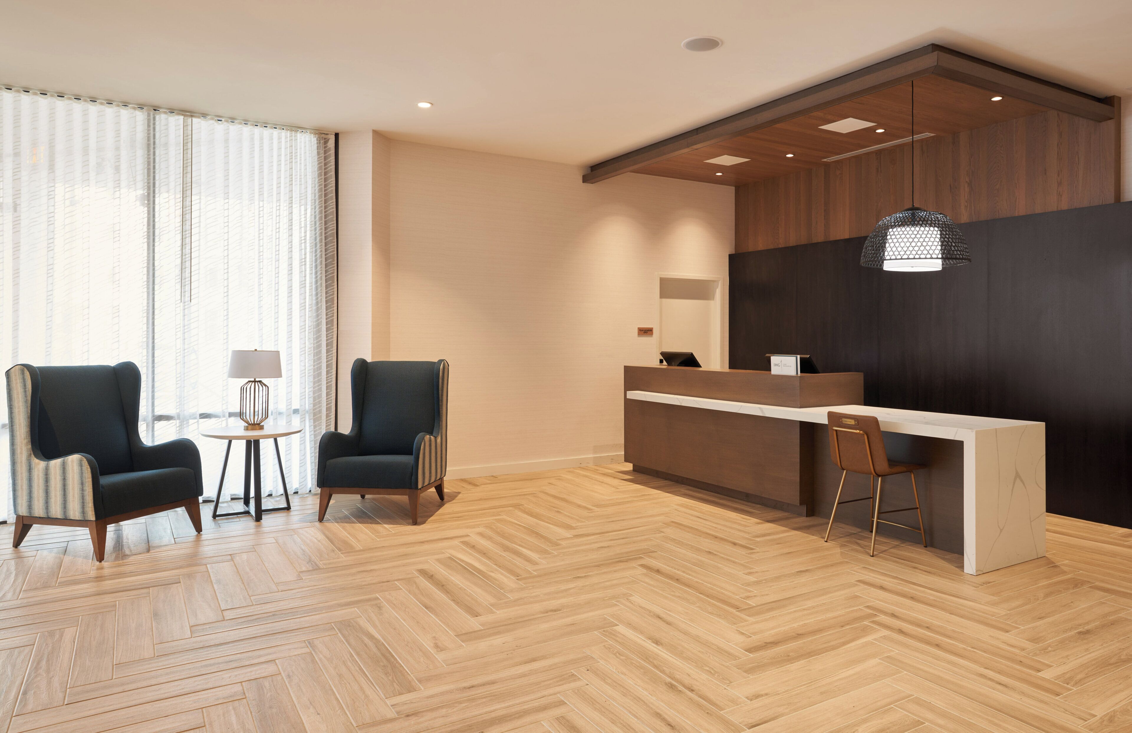 A hotel reception area with a wooden herringbone floor, two armchairs, a small round table, and a reception desk.