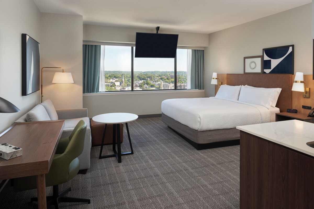 A hotel room with a bed, a desk, a chair, a small table, and a TV.