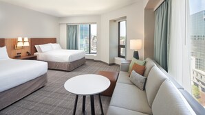 Habitación de hotel con dos camas, un sofá, una mesita y ventanas grandes con cortinas.