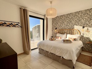 A bedroom with a bed, a wooden headboard, a hanging light fixture, a wall-mounted nightstand, and a window with curtains.