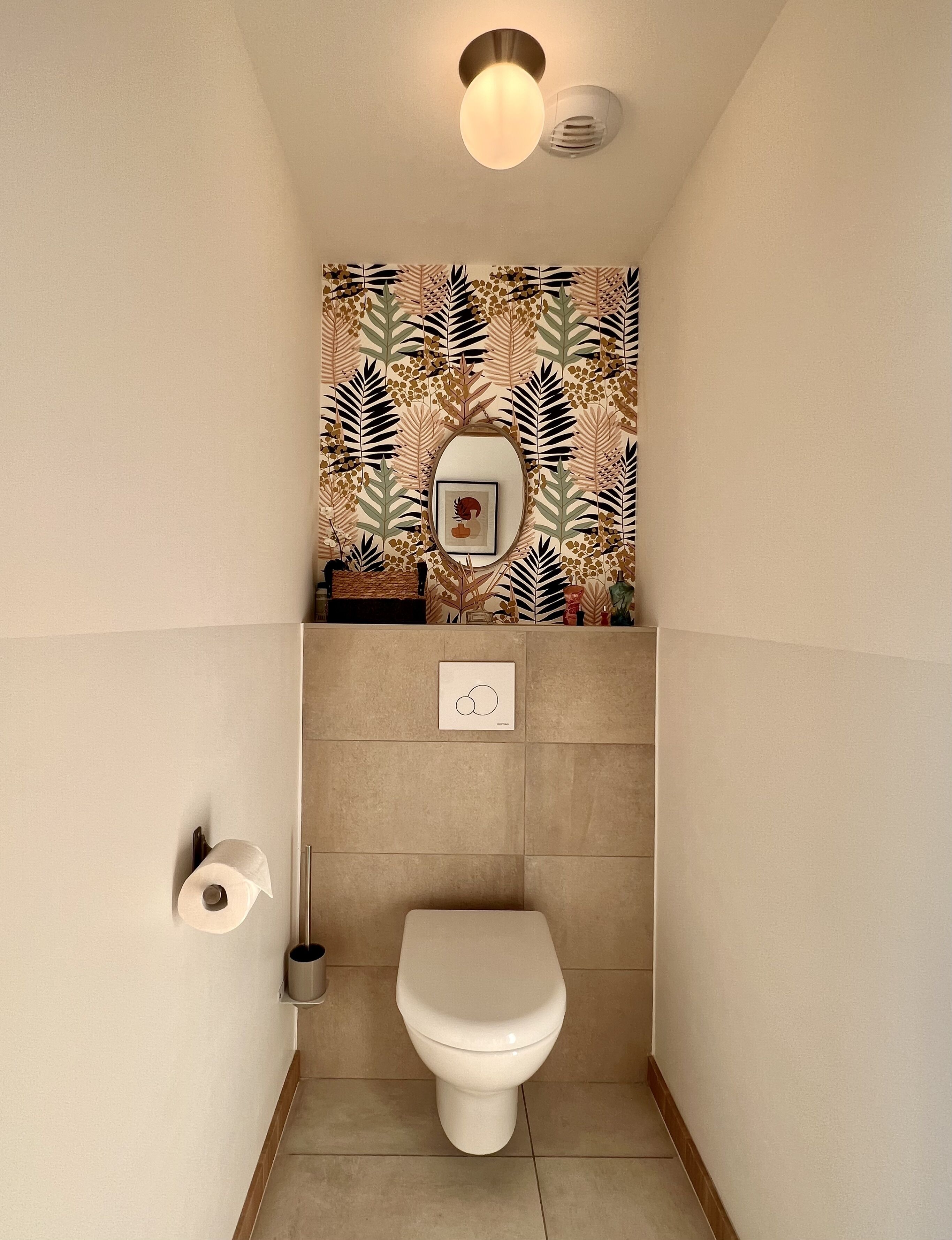 Um banheiro pequeno com vaso sanitário, rolo de papel higiênico fixado na parede, luminária e espelho decorativo.