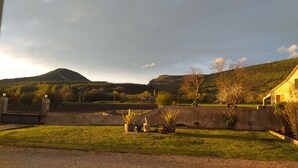 Paisaje rural con una montaña, una zona verde con plantas en macetas y un edificio con techo inclinado.