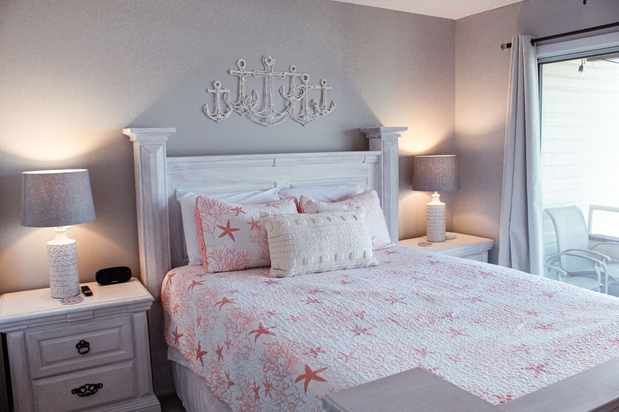 A bedroom with a white bed, two bedside tables with lamps, a decorative wall piece, and a view of the outdoors through a window.