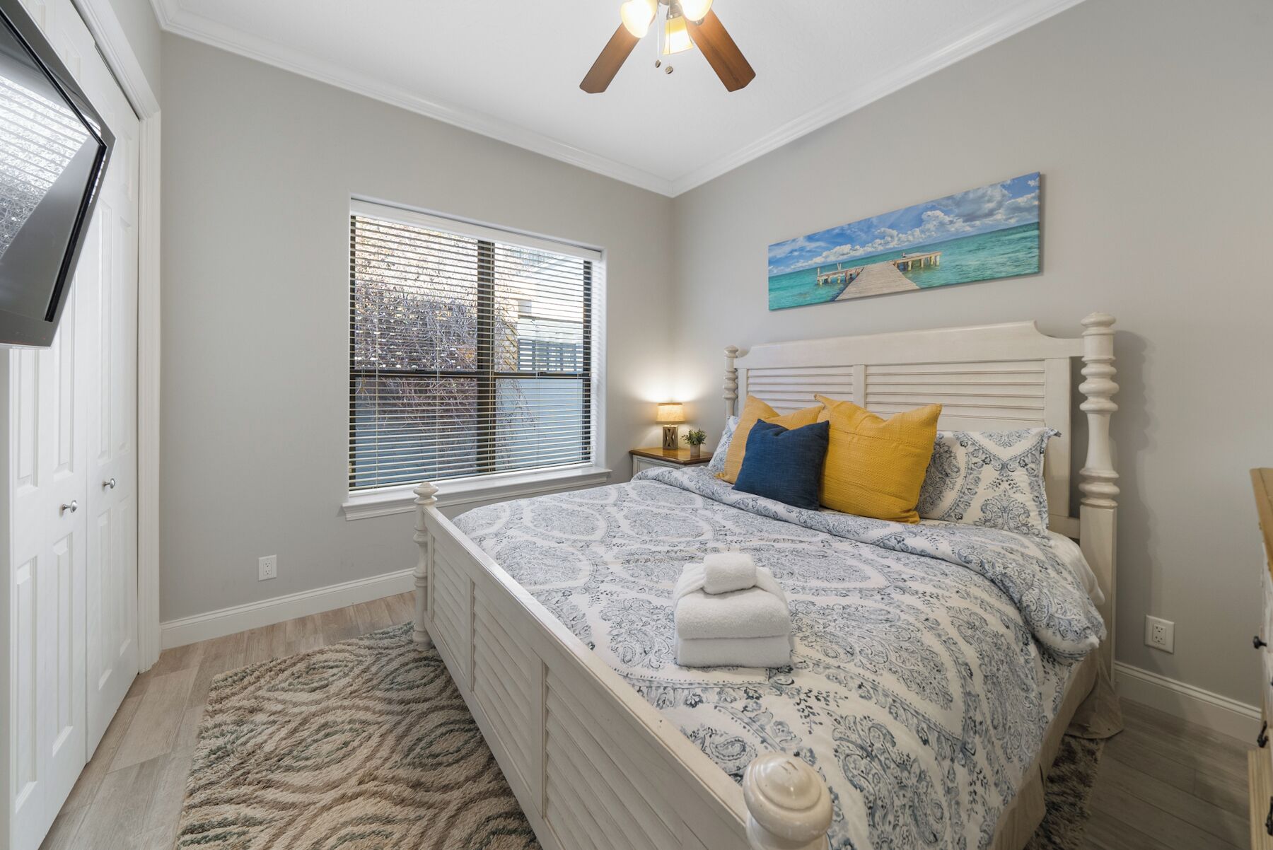 A bedroom with a bed, a window with blinds, a ceiling fan, and a painting of a beach scene.