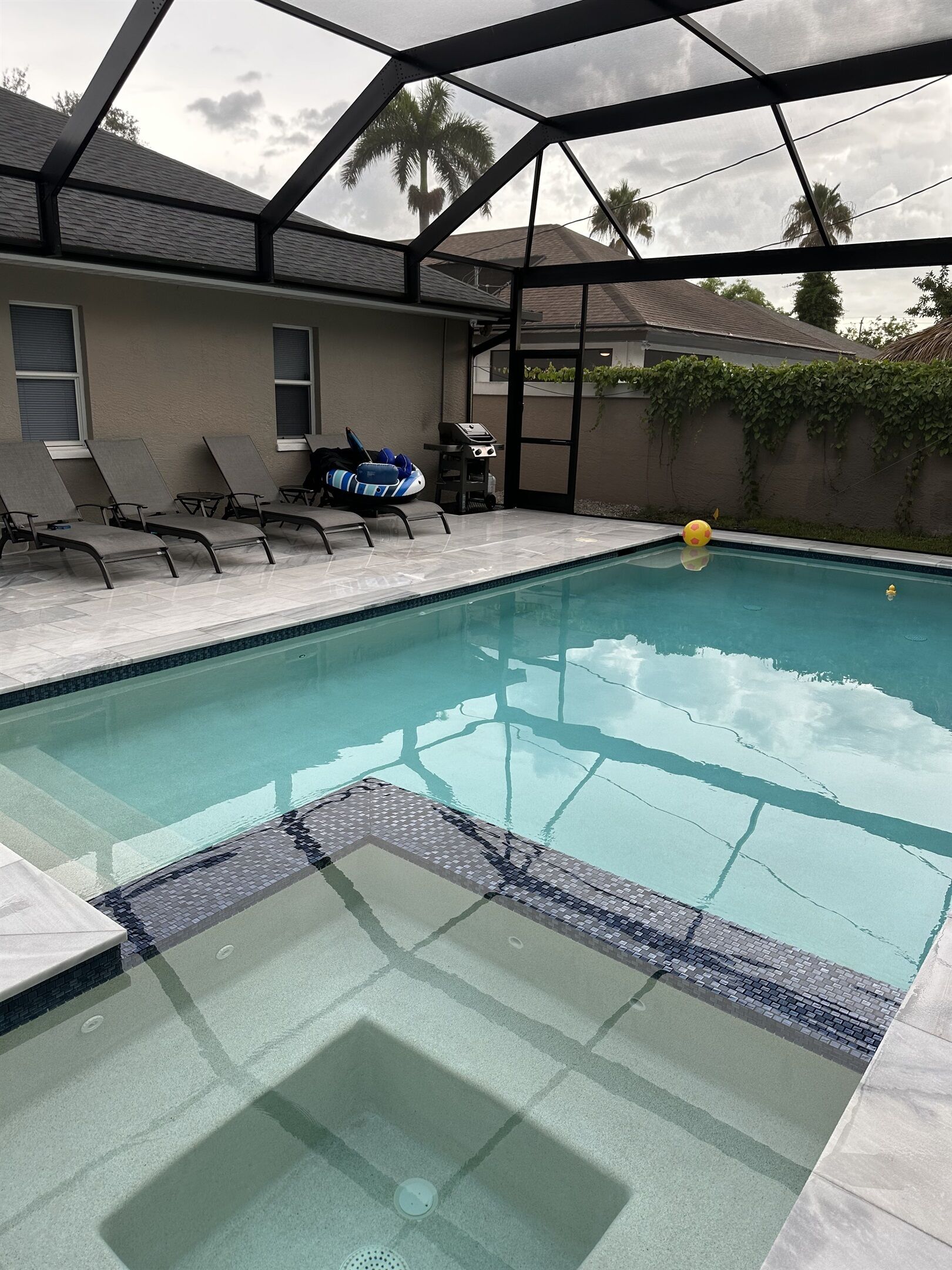 A swimming pool with a glass enclosure, lounge chairs, and a barbecue area.