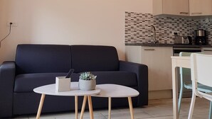 Ett modernt vardagsrum med en mörk soffa, ett runt bord med en växt och ett köksområde med kakel på väggen och träskåp.