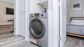 Een kleine, moderne wasruimte met een voorlader wasmachine, een klein plankje met schoonmaakmiddelen, en een bed met een grijze dekbedovertrek in de aangrenzende slaapkamer.