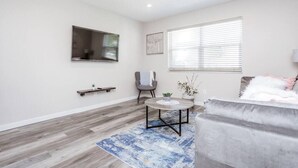 Een moderne woonkamer met een grijze bank, een ronde salontafel, een grijze fauteuil, een blauw vloerkleed, een kleine plant en een raam met jaloezieën.