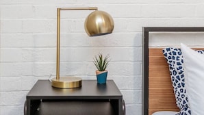 Nachtkastje met een gouden lamp, een kleine potplant en een blauw-wit kussen tegen een witte bakstenen muur.