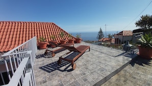 Una terraza en la azotea con suelo de baldosas, plantas en macetas, un banco de madera y vista al mar.