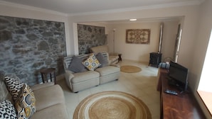 Una sala de estar con una pared de piedra, un sofá color beige, un sillón estampado, una mesa de centro y un mueble de televisión con un control remoto.