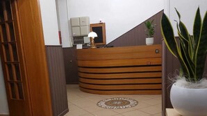A reception area with a wooden reception desk, a tiled floor, a potted plant, and a staircase leading to another floor. - Maison des Roses (Cesenatico)