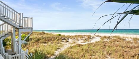 A beach view with a wooden staircase leading to a deck, surrounded by grass and sand.