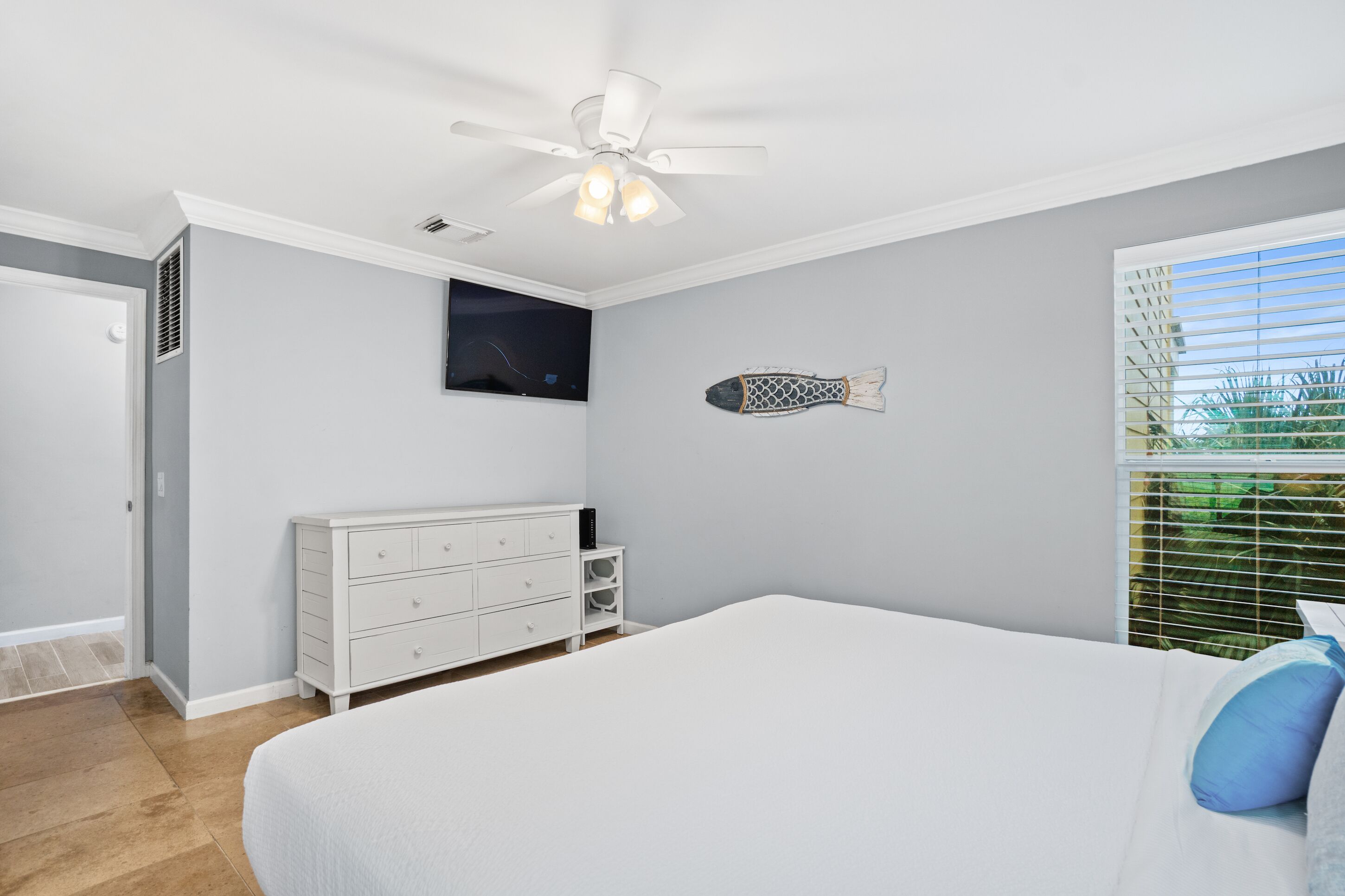 Un dormitorio con cama, cómoda blanca, ventilador de techo, televisión y ventana con persianas.
