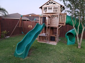 A wooden playhouse with two slides and a small tree in the yard.