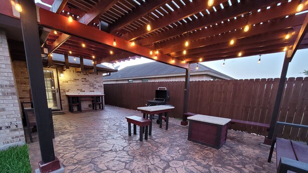 Een terras buiten met stenen vloer, een barbecue en een pergola met lichtjes.